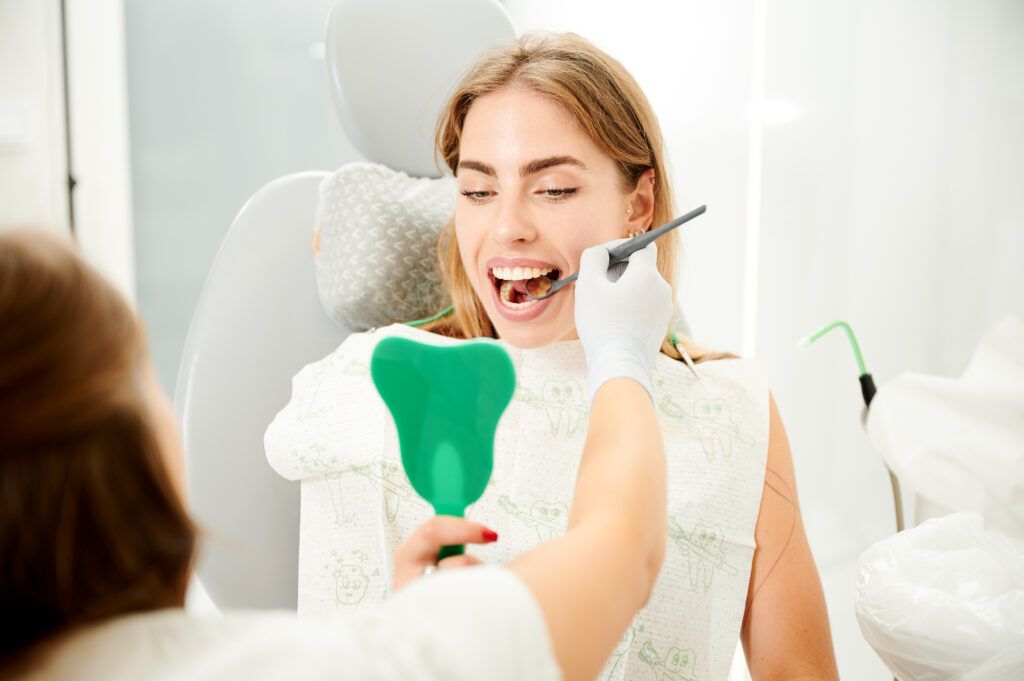 Professional dentist showing female client her sick tooth. Young girl checking up her teeth. Young female looking in mirror on her tooth.