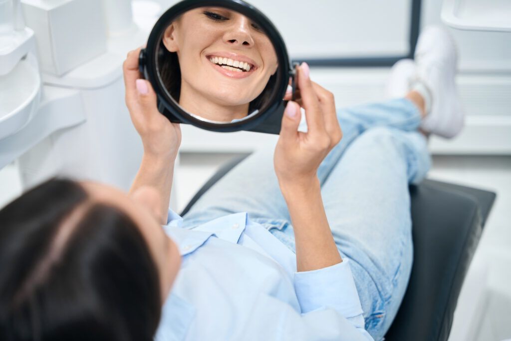 Attractive woman patient lying on orthodontic chair and looking at mirror on her new healthy and white smile, teeth dentures or bridge installation, dental clinic service